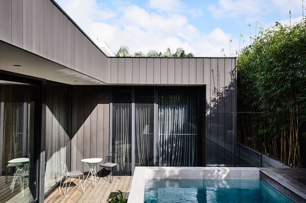 Contemporary single-storey courtyard-style home exterior with dark vertical cladding, extensive glazing, a timber deck, and a compact swimming pool enclosed by dense bamboo planting. The low flat roof and clean geometric lines create a private, modern outdoor living area.