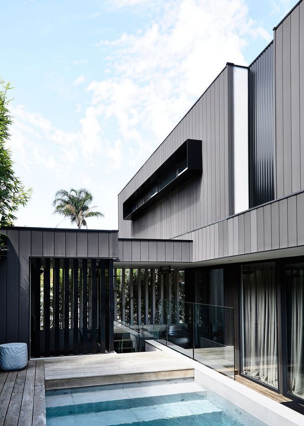 A contemporary multi-level residential courtyard exterior with dark grey vertically ribbed cladding, sharp projecting volumes, narrow clerestory-like windows, and extensive dark-framed glazing. A timber deck surrounds a small rectangular swimming pool, with a frameless glass balustrade, covered outdoor area, vertical privacy screening, and lush planting visible around the edges. The composition is geometric and minimalist, with strong contrast between charcoal facade elements, pale timber decking, and blue pool water.