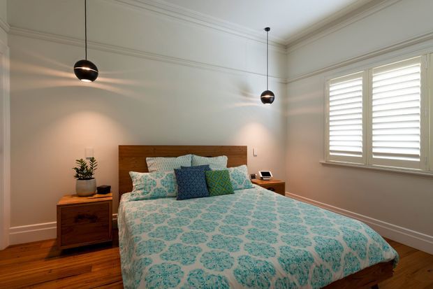 A warm, well-appointed bedroom with a timber bed and matching bedside tables, dressed in a turquoise and white patterned quilt and layered cushions. The room has polished timber flooring, pale walls with decorative ceiling cornices, black pendant lights, and a large window fitted with white plantation shutters.