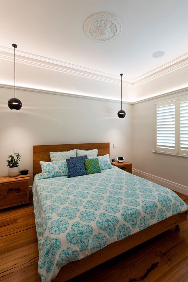 A bright, refined bedroom with a timber platform bed, patterned turquoise-and-white bedding, matching timber bedside furniture, and decorative cushions. The room features warm timber flooring, pale walls, elaborate ceiling cornices with a decorative ceiling medallion, two black globe pendant lights, and white plantation shutters.