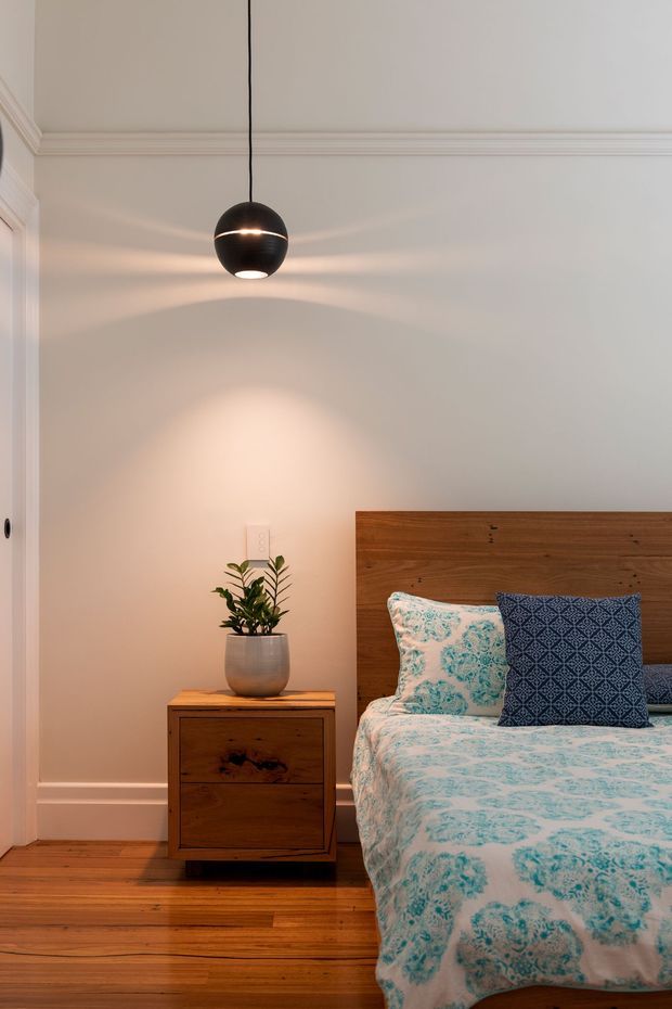 A warmly lit bedroom corner with a wooden bed featuring a tall timber headboard, patterned turquoise-and-white bedding, and a dark blue accent cushion. A matching timber bedside cabinet holds a potted plant, while a black spherical pendant light hangs from the ceiling. White painted walls with decorative crown moulding and polished timber flooring complete the room.
