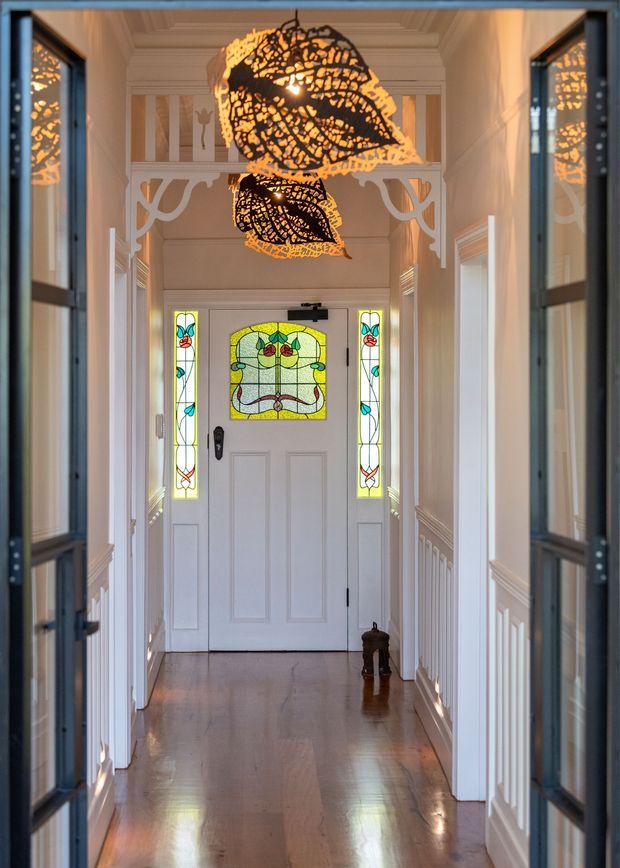 A refined residential entry hall viewed through glazed doors, featuring a polished timber floor, white painted wall panelling and decorative trim, a white front door with colourful stained-glass panels, and striking leaf-shaped pendant lights. The corridor has a traditional, ornate character with symmetrical detailing and warm natural light.