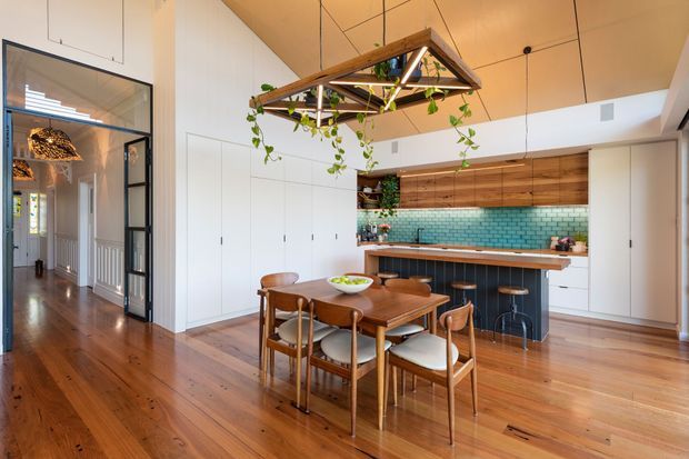 Bright open-plan dining and kitchen area with warm timber flooring, a large wooden dining table and mid-century style chairs, white built-in cabinetry, and a compact kitchen featuring dark lower cabinetry, timber shelving, and a turquoise subway-tile splashback. A timber-framed hanging planter/light structure forms a feature above the dining table. The room opens to a hallway through black-framed glazed doors and has a high, angled ceiling.