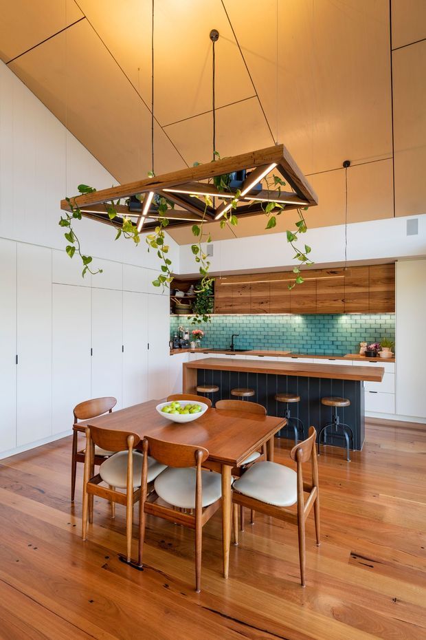 A warm, open-plan kitchen and dining area with a timber dining table and upholstered wooden chairs in the foreground. The kitchen features white floor-to-ceiling cabinetry, a timber island with bar seating, a teal-green subway tile splashback, and timber cabinetry. A distinctive suspended timber-frame pendant light with integrated linear lighting and trailing plants hangs above the dining table. The space has a high sloping timber-panelled ceiling and polished timber flooring.