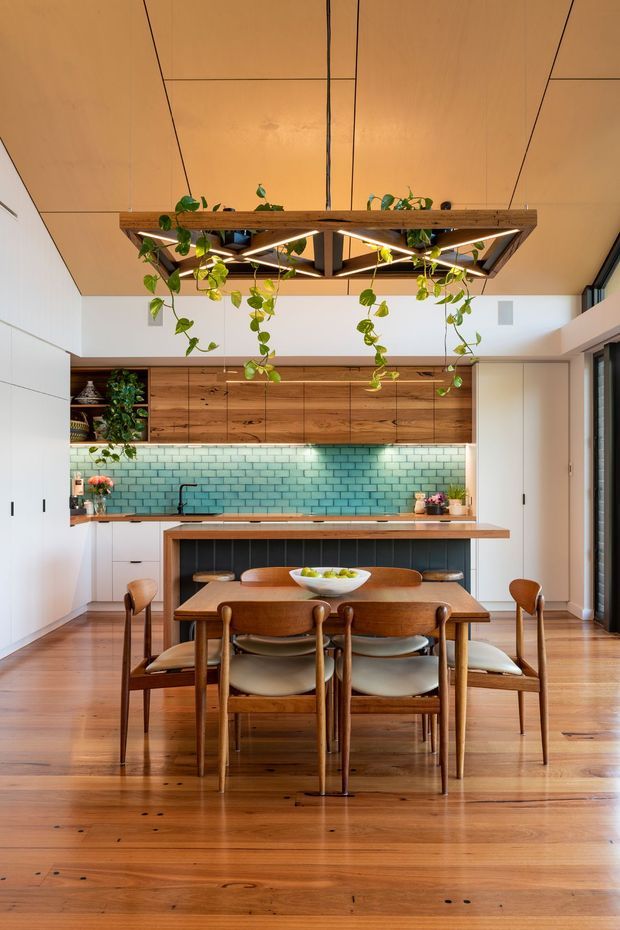 A warm open-plan kitchen and dining area featuring extensive timber cabinetry, a turquoise glazed subway-tile splashback, white integrated storage, and a timber island with a raised dining table. Mid-century-inspired timber dining chairs surround the table, while a suspended timber-framed light fixture with trailing greenery forms a central feature. Polished timber flooring and abundant natural light create a welcoming, refined atmosphere.