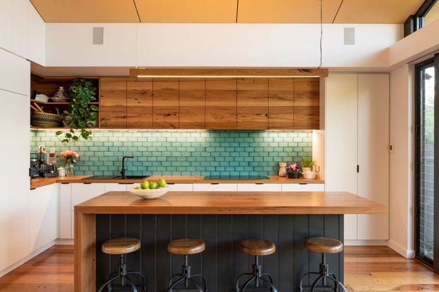 Warm, contemporary kitchen with a large timber island and four stools, extensive timber cabinetry, a mint-green subway-tile splashback, white walls and cabinetry, and clerestory glazing. The open-plan space is naturally lit and features timber flooring, integrated appliances, open shelving with plants and kitchenware, and a dark vertical-panel island base.