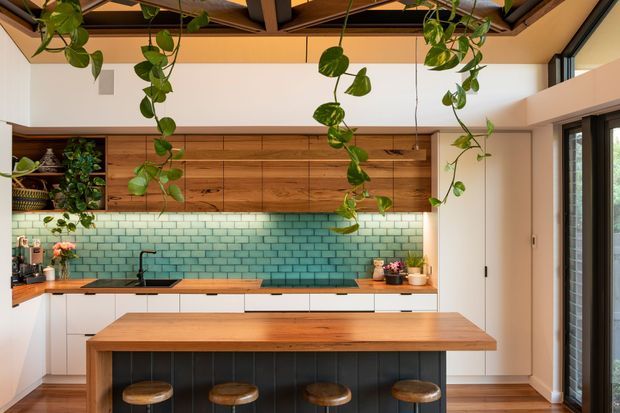 Bright contemporary kitchen with a large timber island, integrated timber cabinetry, white lower cabinets, and a glossy teal-green subway tile splashback. Hanging trailing plants, exposed dark ceiling beams, clerestory glazing, and tall windows create an airy indoor-outdoor feel.