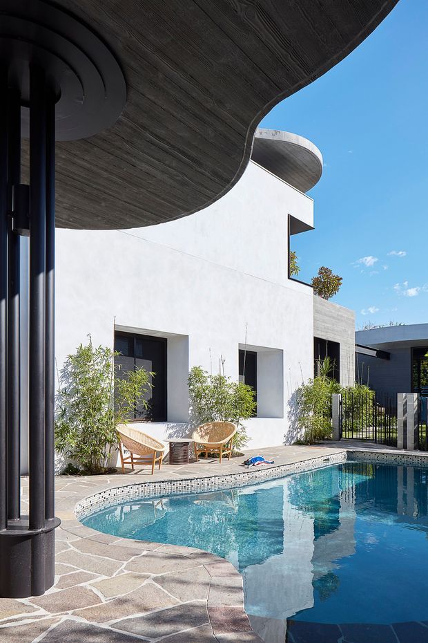 A bright modern courtyard pool area enclosed by white rendered walls and dark architectural elements. The curved swimming pool is edged with small mosaic tiles and irregular stone paving, while sculptural curved roof or canopy forms and cylindrical black columns create a strong contemporary composition. Outdoor lounge chairs sit beside the pool, with planted screening and a clear blue sky beyond.