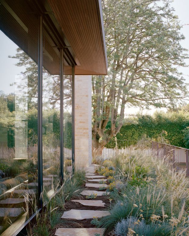 Exterior architectural view of a contemporary house surrounded by a naturalistic landscaped garden. Full-height dark-framed glazing reflects the garden, while a projecting roof with warm timber soffit extends over a pale brick or stone-clad wall. Irregular stepping stones lead through ornamental grasses and low planting toward a timber-railed edge, with mature trees forming the backdrop.
