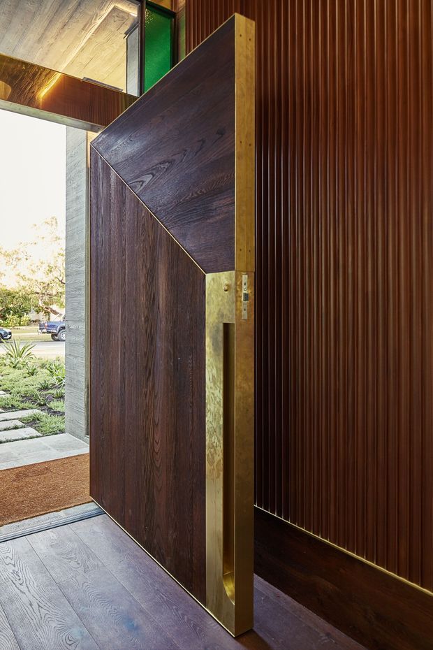 Close-up view of a dramatic contemporary entryway with a tall pivot front door in dark timber, angular geometric detailing, and brass-toned edging and hardware. The interior wall is clad in rich vertical timber fluting, complemented by dark timber flooring and a partially visible timber-lined ceiling. The open doorway reveals a landscaped front path and street outside.