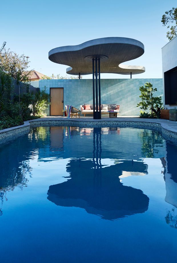 A landscaped residential courtyard centered on a swimming pool, with a sculptural organic concrete canopy supported by slender dark posts over an outdoor lounge. The pool has a tiled edge and reflects the canopy, while a turquoise tiled boundary wall, seating, planting, and part of a modern house frame the scene. Bright daylight and strong mid-century architectural character are evident.
