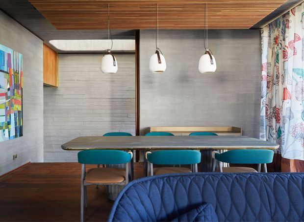A contemporary dining area with a long dark dining table, turquoise upholstered chairs, and a partially visible quilted blue sofa in the foreground. The room features grey concrete-look walls, dark timber flooring, a slatted timber ceiling, wood cabinetry, suspended white pendant lights, and colourful patterned curtains.