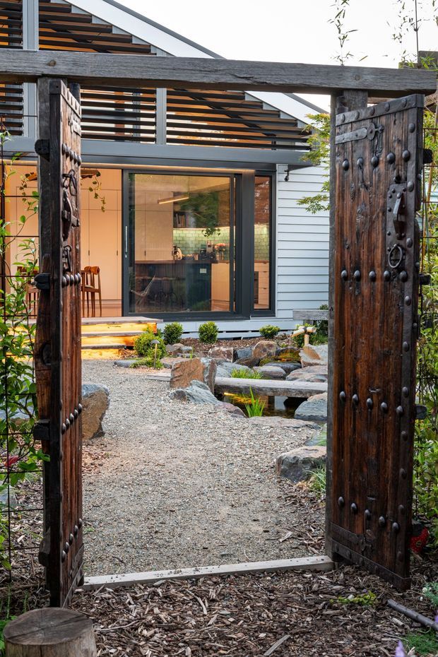 A landscaped courtyard garden viewed through an open pair of substantial weathered timber doors with dark metal studs and hardware. Beyond the gateway is a gravel path, sculptural rocks, small planted beds and a narrow water feature leading toward a contemporary house. The house has dark-framed glazing, horizontal light-coloured cladding, timber screening and a steeply sloping roofline. The scene combines rustic reclaimed materials with contemporary residential architecture.