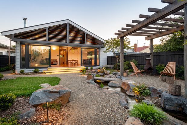 A contemporary single-storey backyard dwelling or extension with a prominent gabled roof, horizontal timber screening beneath the gable, and broad glazed doors opening onto a small deck. The landscaped garden features gravel paths, natural rock edging, a shallow stone-lined water feature, planting beds, and a timber pergola with outdoor seating and a chiminea.