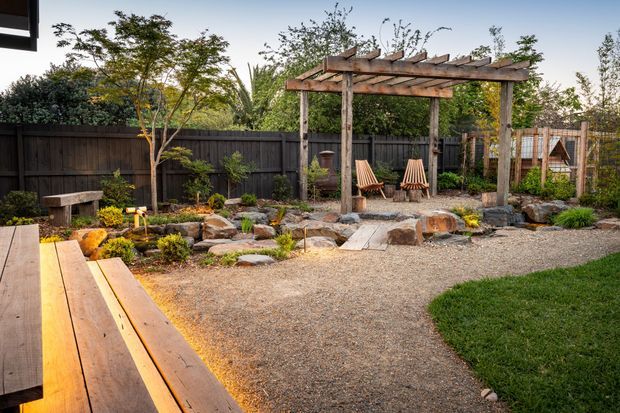 A landscaped backyard garden with a rustic timber pergola, gravel seating area, Adirondack-style timber chairs, natural stone edging and stepping stones, raised timber decking in the foreground, and dense planting enclosed by a dark timber fence.