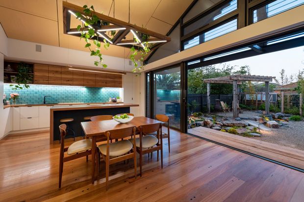 A warm, open-plan kitchen and dining area with timber flooring and cabinetry, a turquoise tiled splashback, and a central island with seating. A timber dining table sits beside large black-framed glazed doors opening onto a landscaped garden courtyard. High-level clerestory glazing and a sculptural timber-and-metal pendant with trailing plants add character and natural light.