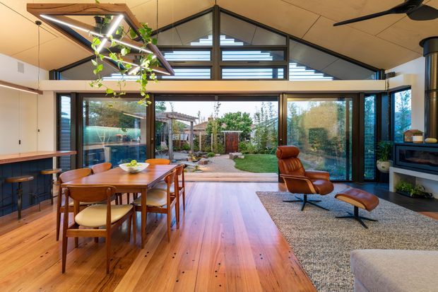 Open-plan contemporary living and dining space with extensive floor-to-ceiling glazing opening to a landscaped courtyard. The room features warm timber flooring, a timber dining table and chairs, a sculptural lounge chair with ottoman, dark-framed windows, a high pitched timber ceiling, clerestory glazing, and a built-in fireplace/media unit. A large suspended timber-framed skylight or light well with trailing greenery adds a distinctive feature overhead.
