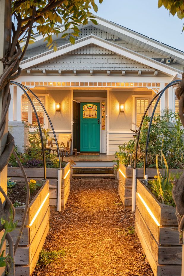 Front exterior view of a charming single-storey home with a prominent gabled porch, white painted weatherboard cladding, decorative gable detailing, and a vivid teal front door with stained-glass panels. A landscaped entry path leads through raised timber garden beds illuminated by integrated linear lighting, with mature trees framing the facade.