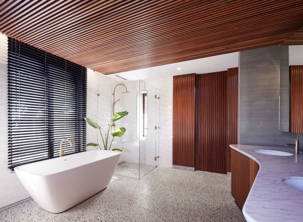 Luxurious contemporary bathroom featuring a freestanding white bathtub, frameless glass shower, double vanity, dark timber slatted ceiling, vertical timber wall panelling, concrete column, terrazzo-style flooring, and black horizontal blinds. The room is bright and spacious with warm timber accents and sculptural curved fixtures.