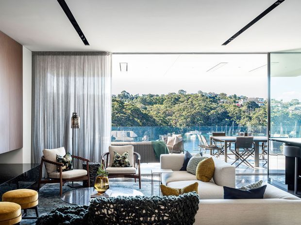 A bright, contemporary open-plan living room with a large cream sectional sofa, sculptural knitted throw, patterned armchairs, glass coffee table and dining area. Full-height glazing opens to a balcony or terrace overlooking a leafy hillside and marina, while black linear ceiling details, timber wall panelling and sheer curtains add architectural definition.