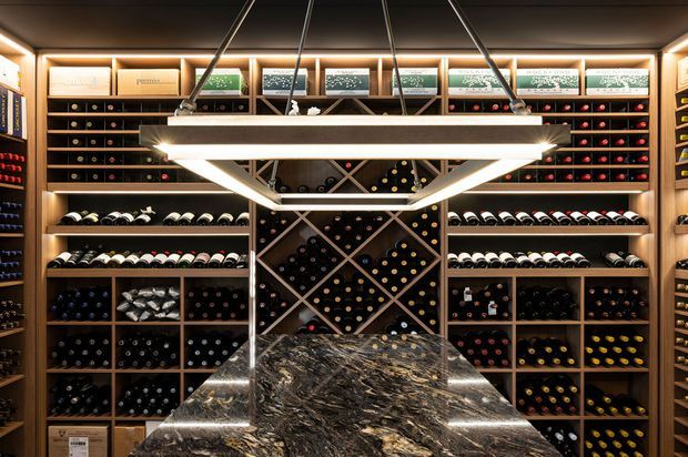 A dramatic, highly organised wine cellar with floor-to-ceiling timber bottle storage covering the walls. Bottles are arranged in horizontal racks and geometric diamond-shaped central display niches, with illuminated label shelves along the top. A large dark patterned stone island or tasting counter occupies the foreground beneath a contemporary suspended geometric pendant light. The space has warm integrated lighting, dark timber cabinetry, and a luxurious, refined atmosphere.