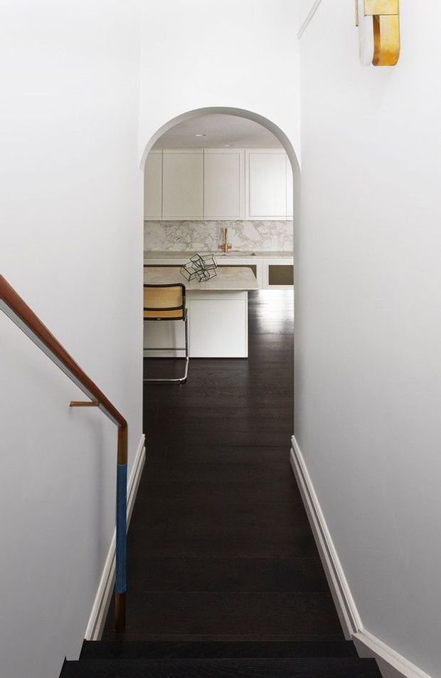 View from a narrow stairway or hallway through a rounded archway into a compact kitchen. The space has white walls and cabinetry, a pale veined stone benchtop and splashback, dark timber flooring, a small island with a metal-framed stool, and a timber handrail with blue detailing. A brass wall light is visible at the upper right.