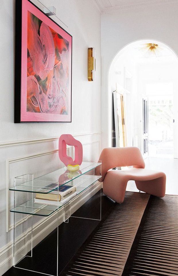 Bright, refined residential entry hall with white decorative wall panelling, a large arched doorway, and a glass console table styled with art objects and books. A sculptural coral-pink lounge chair sits on a dark striped rug beneath colourful abstract artwork and a slim brass wall light.
