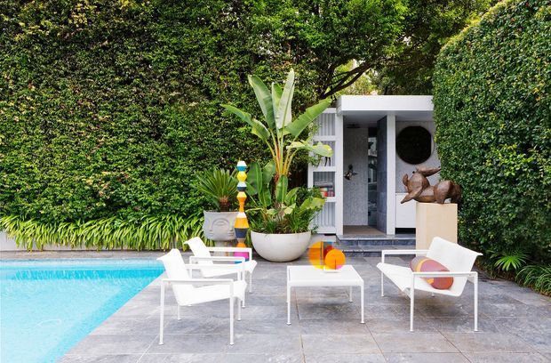 A refined outdoor poolside terrace with pale stone paving, white modern lounge chairs, a low square coffee table, and a vivid turquoise swimming pool. Dense tropical planting and a tall green wall frame a white modern pavilion or pool house with open shelving and sculptural decor. The setting feels lush, bright, and resort-like.