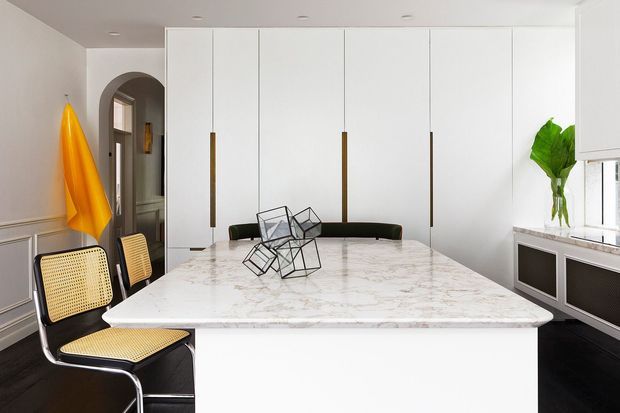 Bright contemporary dining area centered on a large white marble island or dining table, with integrated white cabinetry, slim brass-toned pulls, black seating, and a striking geometric wire sculpture. Traditional wall panelling and an arched opening contrast with the clean modern joinery. A yellow textile and leafy plant provide small colour accents.