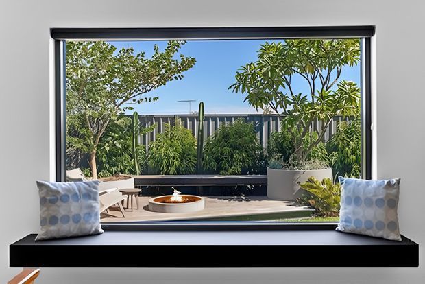 A bright contemporary interior looking through a large black-framed picture window onto a landscaped courtyard or terrace. The deep black built-in window seat has two patterned cushions, while the exterior view includes lush planting, raised planters, a paved area and a low circular fire bowl. The composition emphasises the connection between the indoor seating nook and the garden beyond.