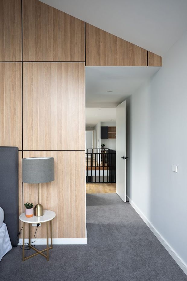 A contemporary bedroom viewed toward an open hallway, featuring a full-height light timber-panelled built-in wall with dark horizontal and vertical reveals. A grey upholstered bed edge, floor lamp, and small round side table sit in the foreground. The room has pale painted walls, grey carpet, recessed ceiling lights, and an open white door leading to a landing with black balustrading and timber flooring beyond.