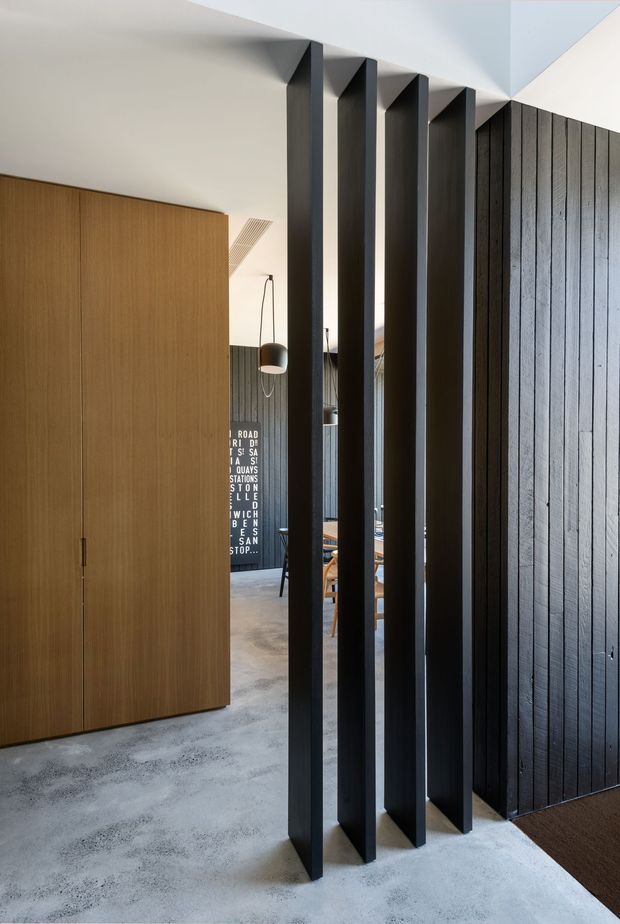 Interior view of a contemporary open-plan space featuring a tall black timber screen with vertical fins, dark vertically grooved wall panelling, a large warm timber built-in cupboard, polished concrete flooring, and a glimpse of dining furniture beyond. The composition emphasizes layered sightlines, strong geometric lines, and contrasting light and dark finishes.