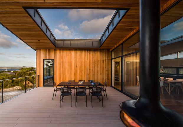 A covered outdoor dining terrace with a timber-lined ceiling and walls, integrated skylight opening, extensive glass sliding doors, and a dark freestanding fireplace in the foreground. A timber dining table and chairs occupy the deck, with a coastal view visible beyond the balustrade. The design has a warm, contemporary indoor-outdoor character with strong architectural timber detailing.