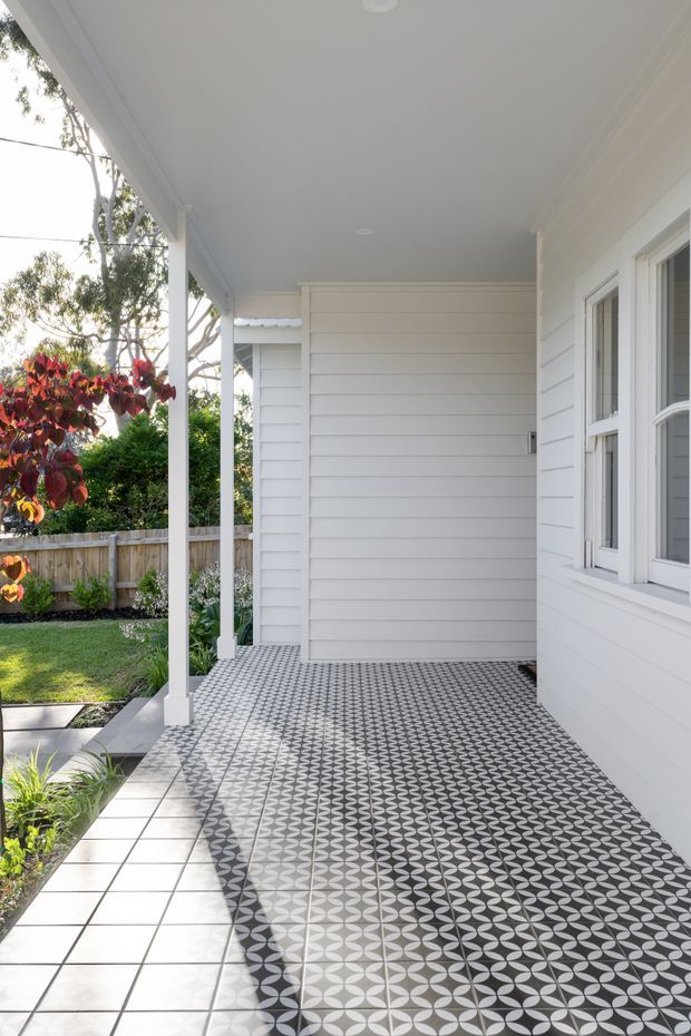A bright covered front porch or verandah with white horizontal weatherboard walls, white posts and trim, and a striking geometric black, white and grey patterned tiled floor. A white-framed window occupies the right wall, while a landscaped garden and timber fence are visible beyond the porch.