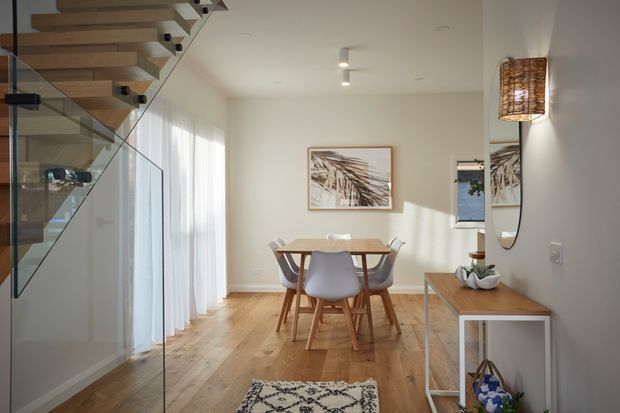 Bright contemporary dining area beneath a floating timber staircase with clear glass balustrading. The room features wide-plank timber flooring, soft off-white walls, sheer curtains, a simple timber dining table with moulded chairs, framed botanical artwork, and a timber console with a woven pendant wall light and large round mirror.