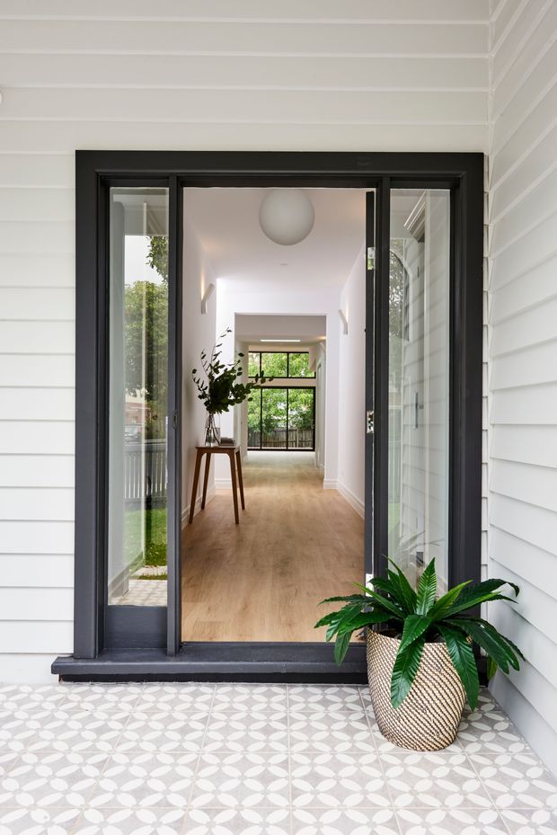 View from a covered front porch through a black-framed glazed entry door into a bright hallway. The exterior has white horizontal weatherboard cladding and patterned light-grey and white encaustic-style porch tiles. Inside, a light timber floor, white walls, a narrow console table with greenery, a spherical pendant light, and full-height glazing at the far end create a clean, airy entrance sequence. A potted plant sits on the porch.