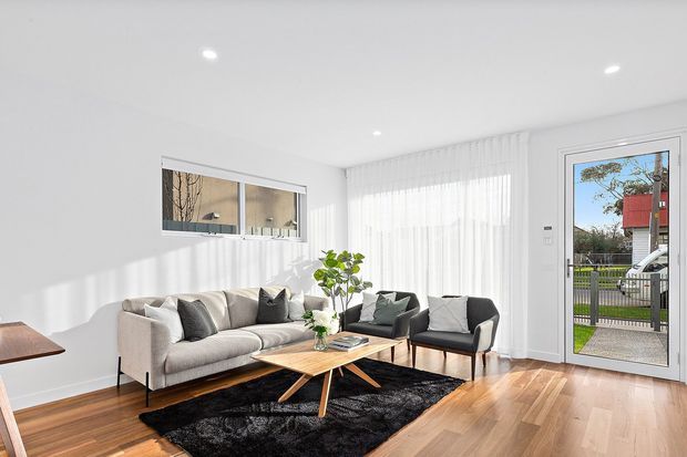 Bright contemporary living room with a grey sofa and matching armchairs arranged around a low timber coffee table on a black rug. White walls, sheer full-height curtains, recessed ceiling lights, timber flooring, and a glazed exterior door create an airy, minimalist interior.