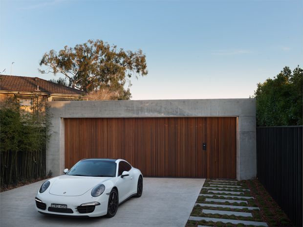 A contemporary residential garage frontage with a broad concrete portal frame and a large vertically slatted timber garage door. A white sports car sits on the smooth concrete driveway, with stepping stones and planted groundcover along one side. Mature trees, a dark fence, and part of a tiled-roof dwelling are visible in the background.
