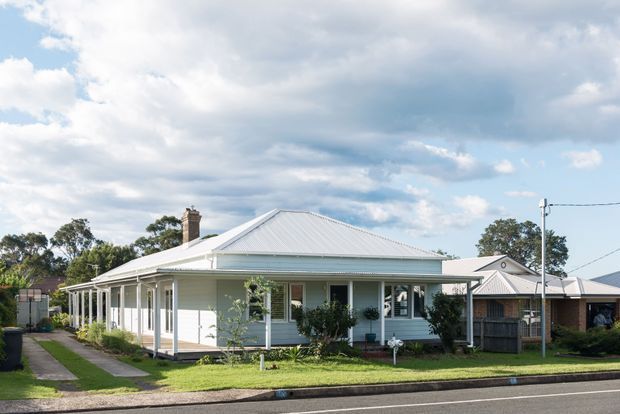 Single-storey detached house viewed from the street, with a broad corrugated hipped roof, pale weatherboard exterior, wraparound veranda with slender posts, chimney, and front garden. The streetscape includes neighbouring low-rise homes and mature trees under a partly cloudy sky.