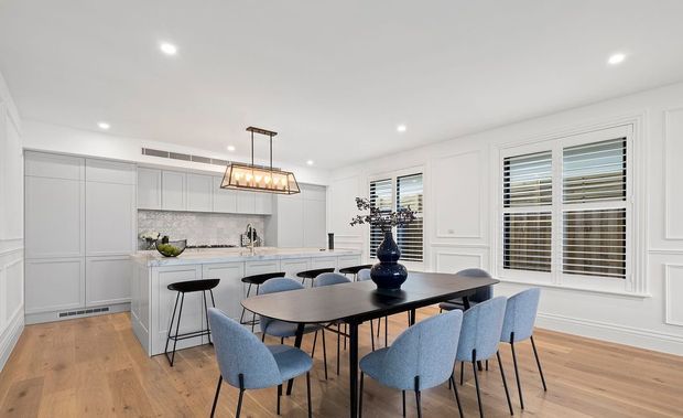 Bright, refined open-plan kitchen and dining area with pale grey shaker-style cabinetry, a light stone benchtop and backsplash, white decorative wall panelling, timber flooring, plantation-shutter windows, and a dark dining table surrounded by upholstered blue-grey chairs. A linear pendant light hangs above the table.