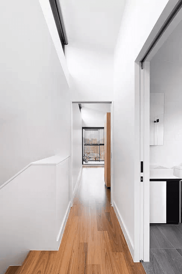 A bright, narrow residential hallway with white walls and trim, warm timber flooring, and a partially visible tiled bathroom or utility room through a sliding doorway on the right. A stair balustrade and open doorway lead toward a rear area with dark-framed glazing and vertical screening. The space has a clean contemporary minimalist appearance with strong linear geometry.