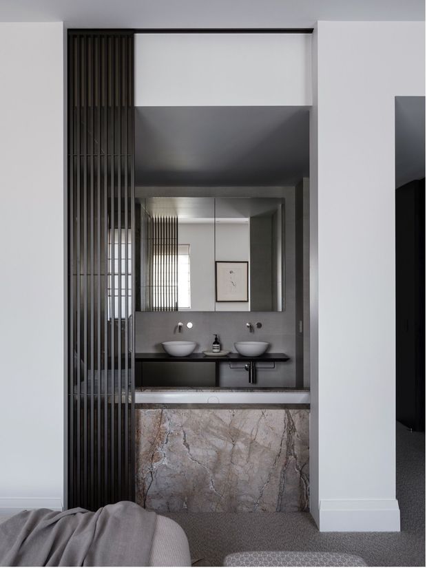 A refined ensuite bathroom viewed directly from a bedroom, with two white vessel basins on a dark vanity, wall-mounted taps, a large mirror, grey wall finishes, and a dramatic veined natural-stone front panel. A dark vertical slatted screen partially separates the bathroom from the bedroom, while carpet and bedding are visible in the foreground.