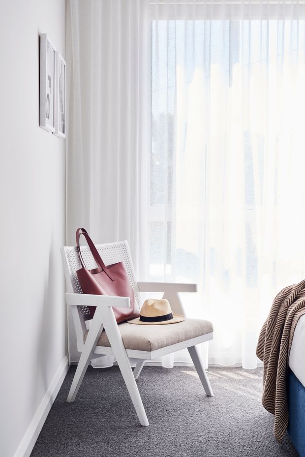 Bright, softly styled bedroom corner featuring a white mid-century-inspired lounge chair with a neutral upholstered seat, a burgundy leather handbag and straw hat, sheer white curtains over a large window, grey carpet, and the edge of a bed with a blue base and textured throw. The room has a clean, airy, minimalist aesthetic.