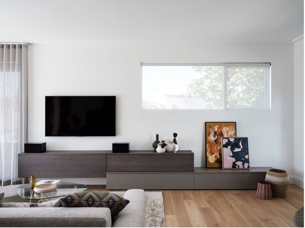Bright contemporary living room with a wall-mounted television above a long dark wood media console, light timber flooring, a pale sectional sofa, patterned cushions and rug, framed artwork, decorative objects, and a large horizontal window with sheer curtains.