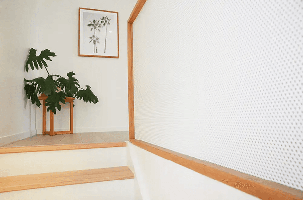 Bright minimalist stairway with pale timber stair treads and trim, white walls, and a textured white perforated wall panel or screen along the stair. A large mirror reflects a leafy indoor plant and a small timber console, while a framed botanical print adds a subtle natural accent.