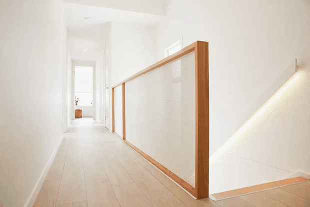Bright minimalist upper-level hallway with pale timber flooring, white walls, and a stair opening protected by a timber-framed translucent or textured glass balustrade. The corridor is softly illuminated with recessed lighting and natural light, creating a clean, airy feel.