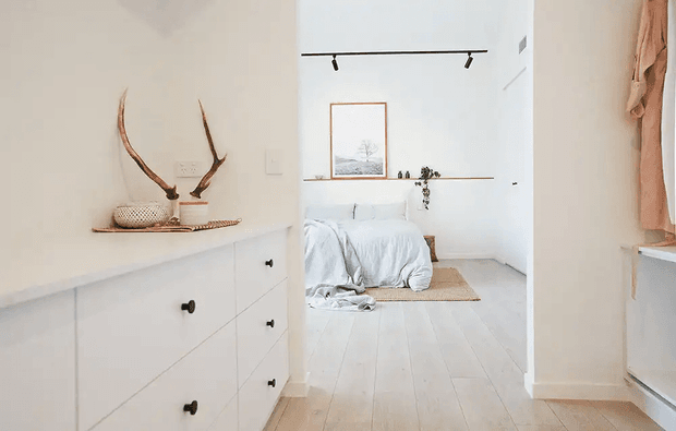 Bright minimalist bedroom viewed through an open doorway, with white bedding, pale timber flooring, built-in white cabinetry in the foreground, a narrow floating shelf, simple framed artwork, track lighting, and natural decorative accents including antlers and woven objects.