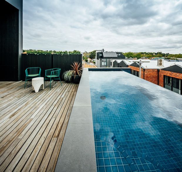 Rooftop terrace with a long blue-tiled swimming pool, timber decking, outdoor chairs and a small table. The contemporary setting overlooks neighbouring low-rise buildings and is framed by dark vertical cladding, planted screening and a cloudy sky.