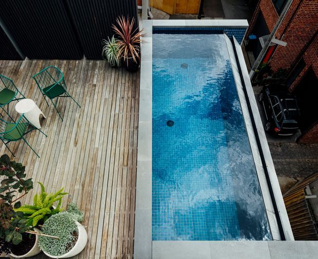 Overhead view of a compact rooftop or elevated outdoor entertaining area with a long rectangular swimming pool finished in small blue mosaic tiles. A timber deck with green outdoor chairs and a small white table runs alongside the pool, with potted plants adding greenery. Adjacent brick walls, fencing, and a parked car are visible around the perimeter.