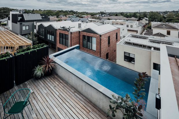 Elevated view across a contemporary urban residential development, featuring a rooftop timber deck and a narrow elevated swimming pool bordered by concrete walls. Surrounding homes combine exposed red brick, dark metal cladding, white rendered facades, and low-pitched metal roofs.